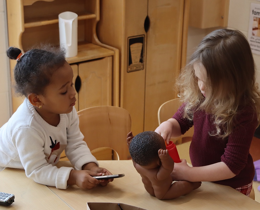 Two young girls in a PLASP Early Learning and Child Care Centre stand at a table together, taking care of a baby doll